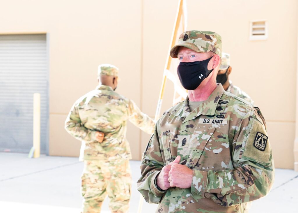 US Army soldiers in camouflage uniforms wearing masks outdoors during the day.
