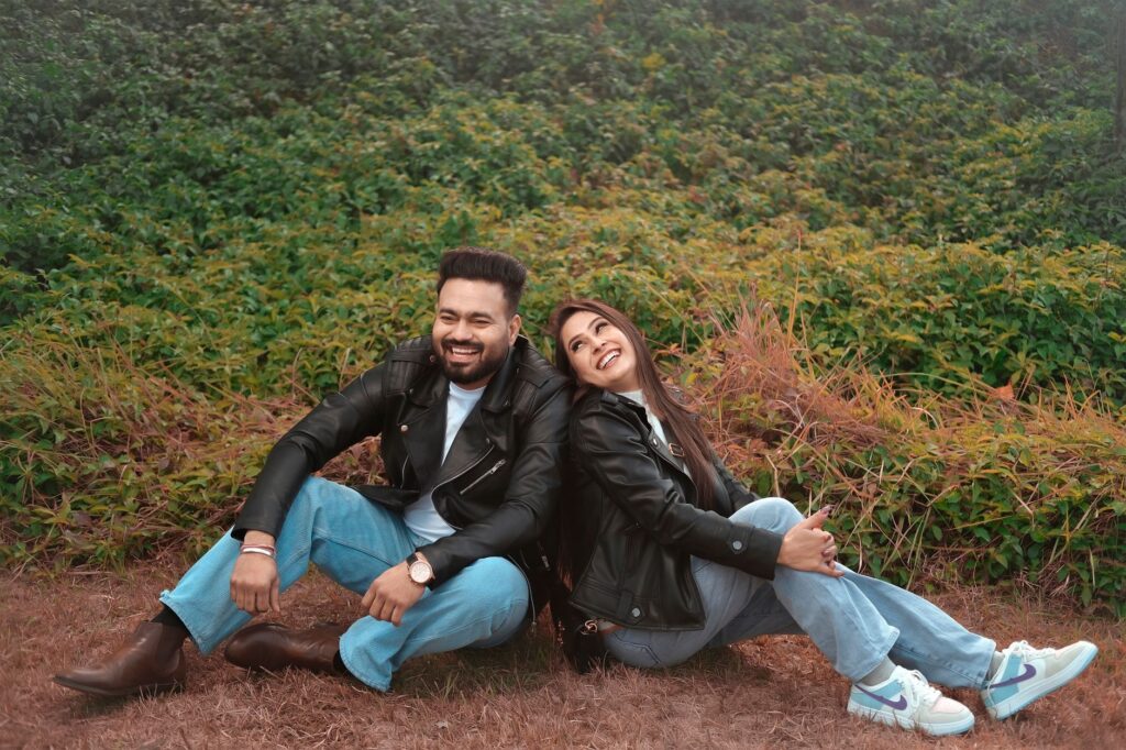 Smiling couple in leather jackets and jeans sitting outdoors in a lush green setting.