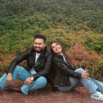 Smiling couple in leather jackets and jeans sitting outdoors in a lush green setting.