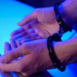 Close-up image of hands in handcuffs resting on a table under blue lighting depicting law enforcement.