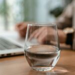 Close-up of a glass of water on a desk in an office setting with a laptop in the background.