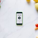 Mobile delivery app surrounded by fresh fruits and vegetables on a marble countertop.